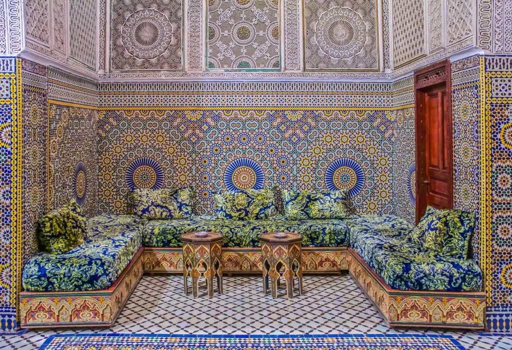 Salon marocain traditionnel orné de zellige bleu, jaune et blanc, avec banquettes recouvertes de tissu fleuri et tables basses sculptées.